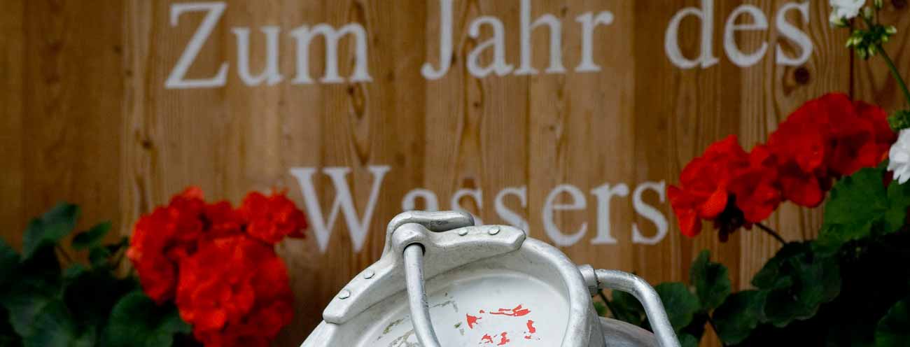 Wasserkanister aus Holz mit Schrift geschmückt mit roten Geranien 